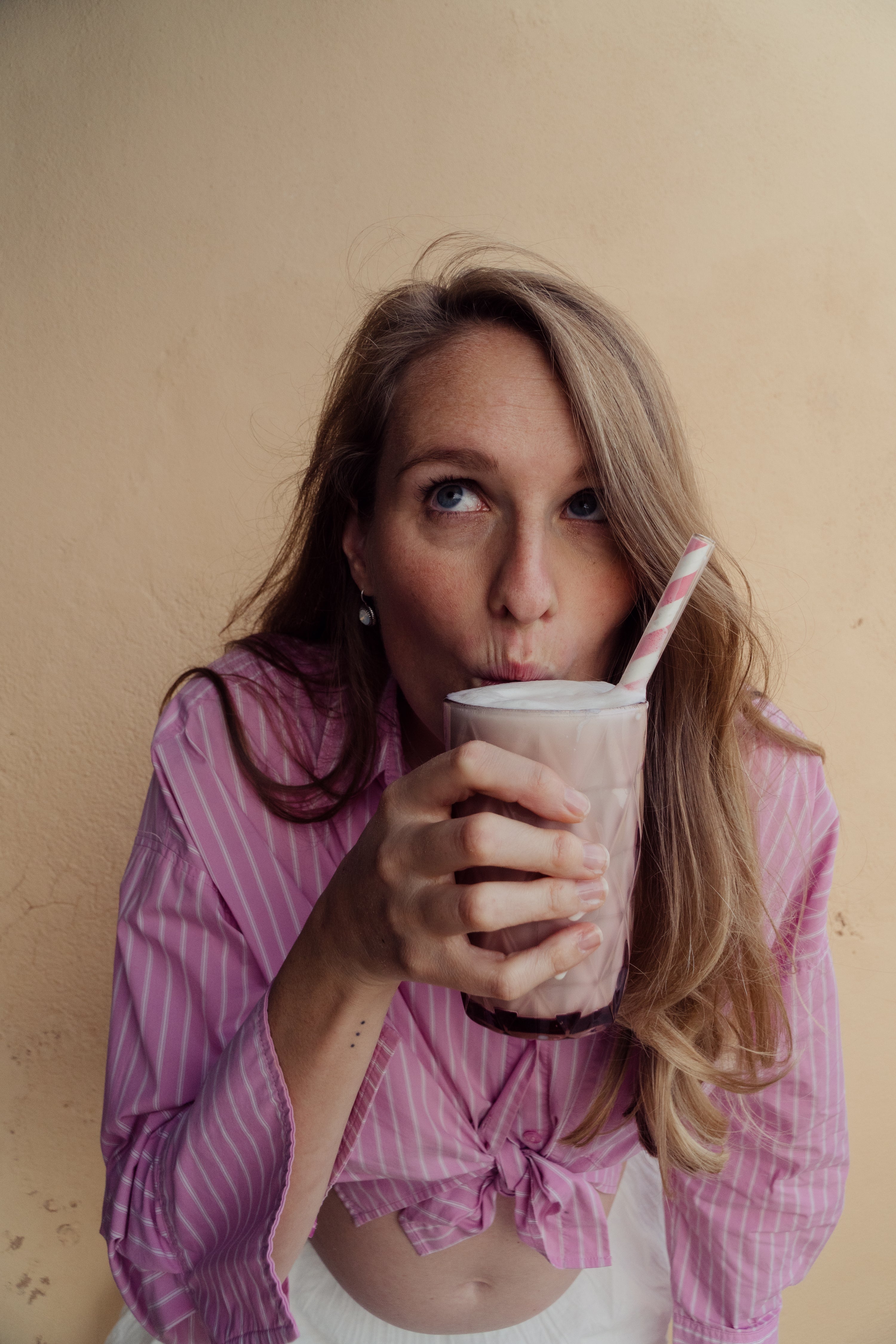 Woman drinking the MomShake from a pink cup with a straw against a beige background on Ibiza